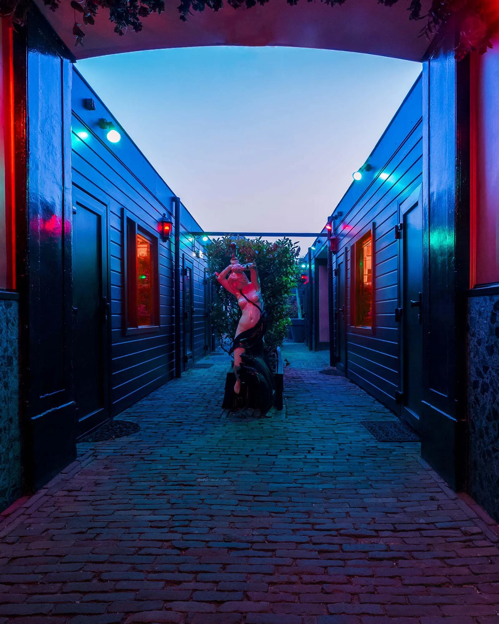 De doorgang tussen de kamers, uitgelicht in rood, groen en blauw, met aan het eind een beeld van een vrouwfiguur tussen de beplanting.