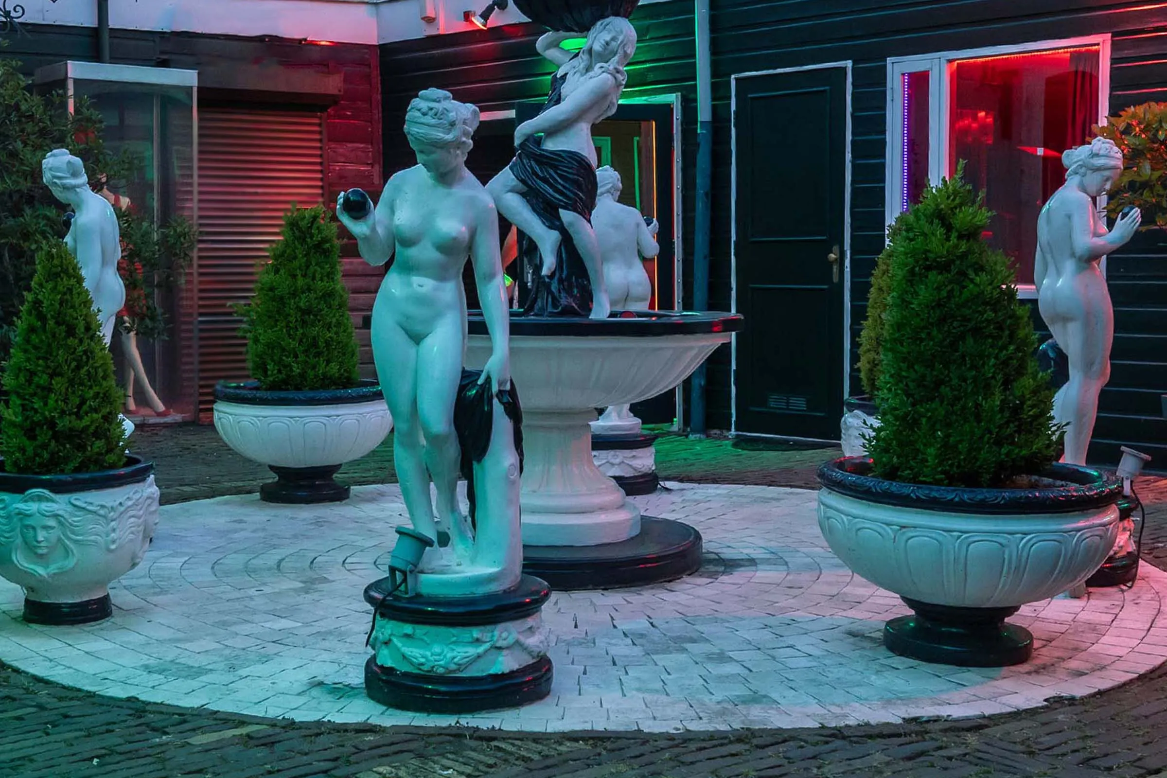 De binnenplaats in de avond, met klassieke witte beelden rond de fontein en de deuren van de kamers in rood en groen licht.