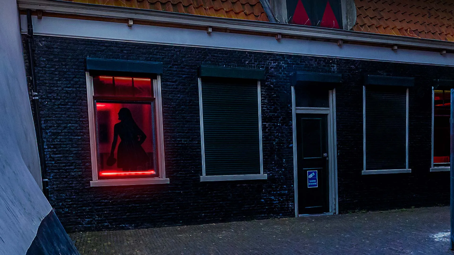 De gevel aan de buitenkant van het pand in de avond, met dichte rolluiken en één rood verlicht raam waarachter het silhouet van een dame staat.
